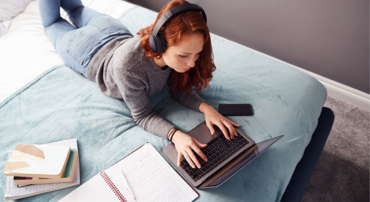 Étudiante sur son lit qui travail sur son ordinateur et des manuels autour